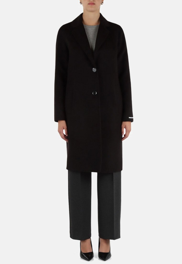 Cappotto di lana nera con chiusura a un pulsante, revers a punta e due tasche laterali, abbinato a pantaloni grigi sartoriali e tacchi neri.