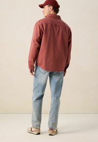 Rode katoenen shirt met lange mouwen, ademende pasvorm en knoopmanchetten, gecombineerd met lichtblauwe jeans en beige sneakers, gezien vanuit de achterkant.