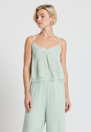 Femme aux cheveux blonds courts portant un caraco vert clair orné de dentelle et un pantalon large assorti, avec de longues boucles d'oreilles pendantes.