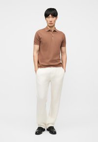 Jeune homme debout les mains dans les poches, portant un polo marron à manches courtes, un pantalon blanc et des chaussures habillées noires sur un fond uni.