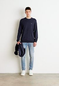 Jūrų pajėgų megztinis, šviesiai mėlynos džinsai ir balti sportbačiai. Asmuo laiko tamsią striukę. Paprastas dizainas, tvirtas kirpimas ir sklandi tekstūra.