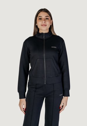 Black zip-up jacket with a high collar, front pockets, and embroidered "GUESS" logo. Made of smooth, stretchy fabric, paired with matching pants.