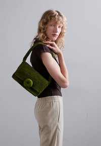 Borsa a tracolla verde intrecciata con forma rettangolare, caratterizzata da una texture morbida e un dettaglio a bottone rotondo. Il modello indossa un top nero e pantaloni beige.