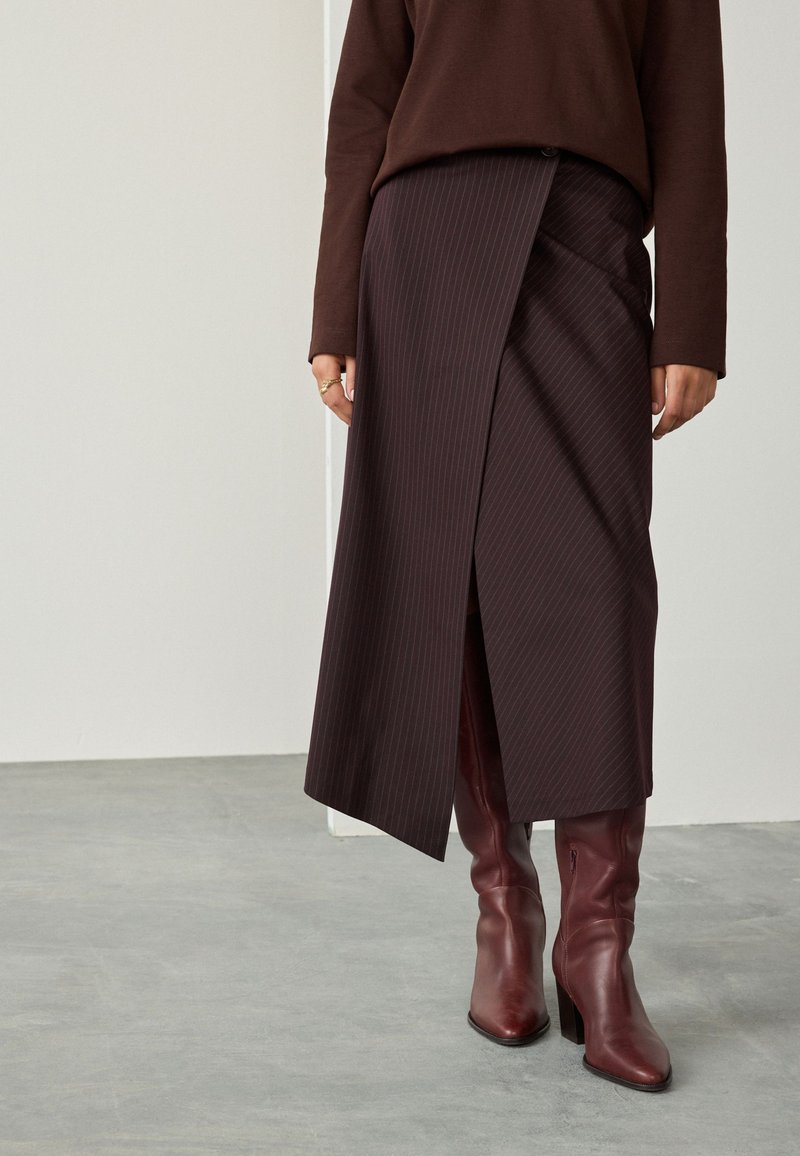 Jupe à rayures fines brun foncé avec un design portefeuille, comprenant une fente latérale et un détail bouton, associée à des bottes hautes bordeaux.