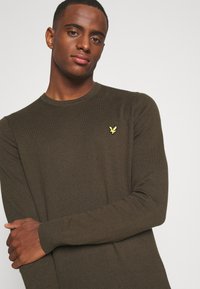 Maglione lungo verde scuro con texture a coste, caratterizzato da un piccolo logo giallo sul petto. Collo rotondo e maniche aderenti.
