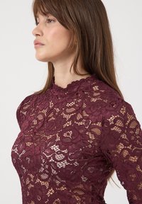 Femme aux longs cheveux bruns portant un haut en dentelle bordeaux à col montant et manches longues avec des motifs floraux, tournée vers la gauche sur fond blanc.
