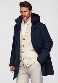 Marineblauwe geïsoleerde parka met een capuchon, met een ritssluiting aan de voorkant, trekkoorden en twee zijzakken, gelaagd over een crèmekleurige gebreide vest.