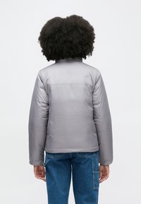 Γκρι μπουφάν με επένδυση και ψηλό γιακά, από λείο ύφασμα με λογότυπο στην πλάτη. Συνδυάζεται με μπλε τζιν παντελόνι για casual εμφάνιση.