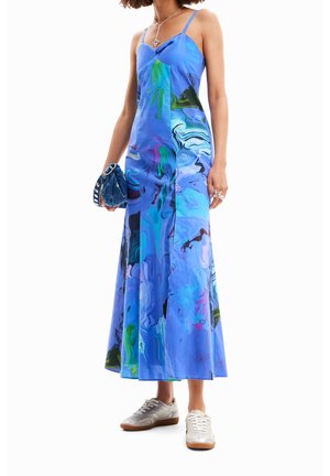 Vrouw draagt een blauwe mouwloze maxi-jurk met abstracte print, zilveren sneakers, zilveren ringen, een hangerketting en houdt een blauwe handtas vast.