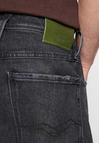 Jeans en denim gris foncé avec une texture rugueuse, présentant une pièce en cuir verte étiquetée "REPLAY" à l'arrière. Coutures apparentes sur les poches.