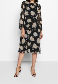 Svart blommig klänning med långa genomskinliga ärmar, prydd med ljusa blommor och rak skärning. Matchad med svarta ankelsandaler.