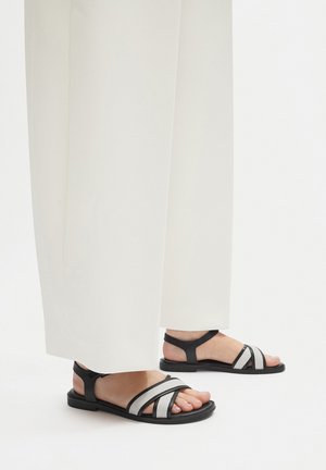 Černé a šedé sandály s pásky, které mají několik pásků a plochou podrážku, doplněné širokými bílými kalhotami.