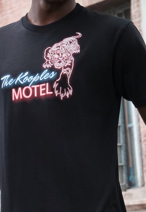 Osoba nosząca czarną koszulkę z grafiką tygrysa w stylu neonowym i napisem "The Kooples MOTEL" na klatce piersiowej.