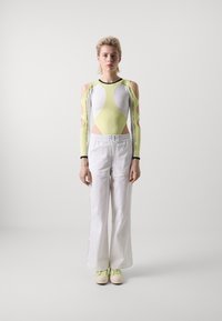 Strakke bodysuit in wit en neongeel met uitsneden bij de armen; gecombineerd met losse witte broeken en lichtgele sneakers met groene veters.