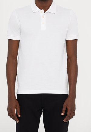 Homme portant un polo blanc uni à manches courtes avec une patte à trois boutons et un pantalon noir, debout, les bras détendus.
