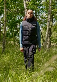 Μαύρο γιλέκο με επένδυση πάνω από ελαφρύ μπλε μακρυμάνικο πουκάμισο, σε συνδυασμό με μαύρο παντελόνι, περπατώντας σε ψηλό πράσινο χορτάρι ανάμεσα σε δέντρα.