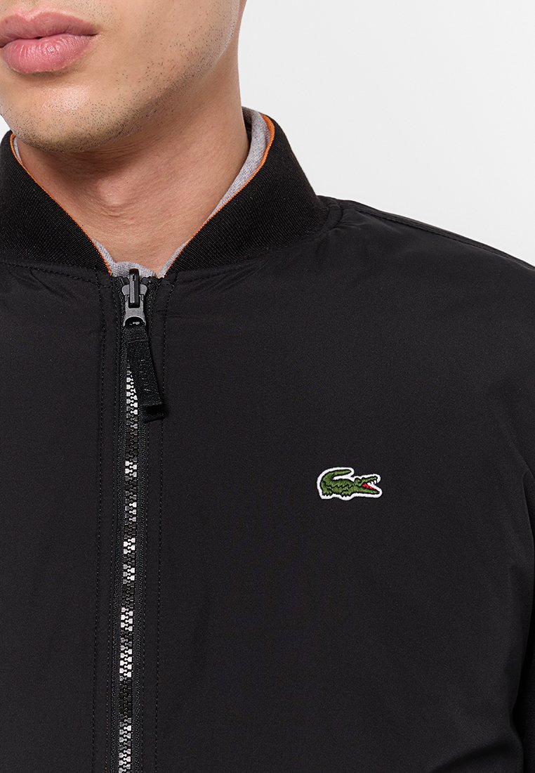 Veste bomber noire avec fermeture éclair, tissu texturé et logo crocodile Lacoste vert sur la poitrine. Accents orange sur le col.