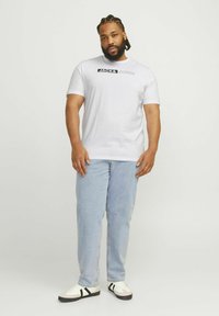 Camiseta blanca de manga corta con un logo negro de "JACK & JONES". Combinada con jeans de color azul claro y zapatillas blancas con franjas negras.