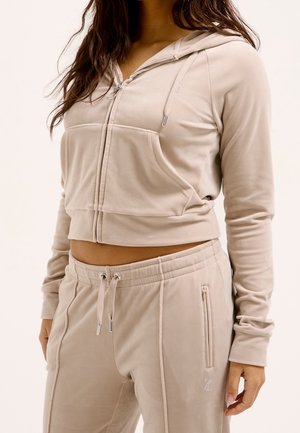 Vrouw draagt beige hoodie met ritssluiting en bijpassende sweatpants met trekkoord, toont blote buik, handen ontspannen langs het lichaam.