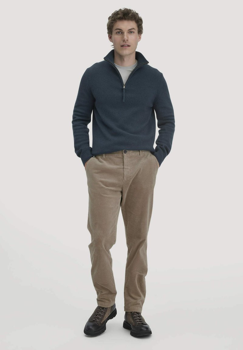 Marine ritsweater, beige corduroy broek en donkere laarzen. De sweater heeft een gestructureerde kraag en de broek heeft een ribbelstructuur.