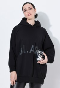 Zwarte oversized hoodie met een glanzende "BLACK" print op de voorkant. Gecombineerd met een kleine metalen zilveren tas. Soepele stoftextuur.