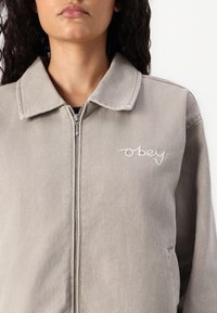 Pilkos džinsinės striukės su užtrauktuku priekyje, smailiu apykakle ir išsiuvinėtu "obey" logotipu kairėje krūtinės pusėje. Matoma tekstūruota medžiaga ir šoninės kišenės.