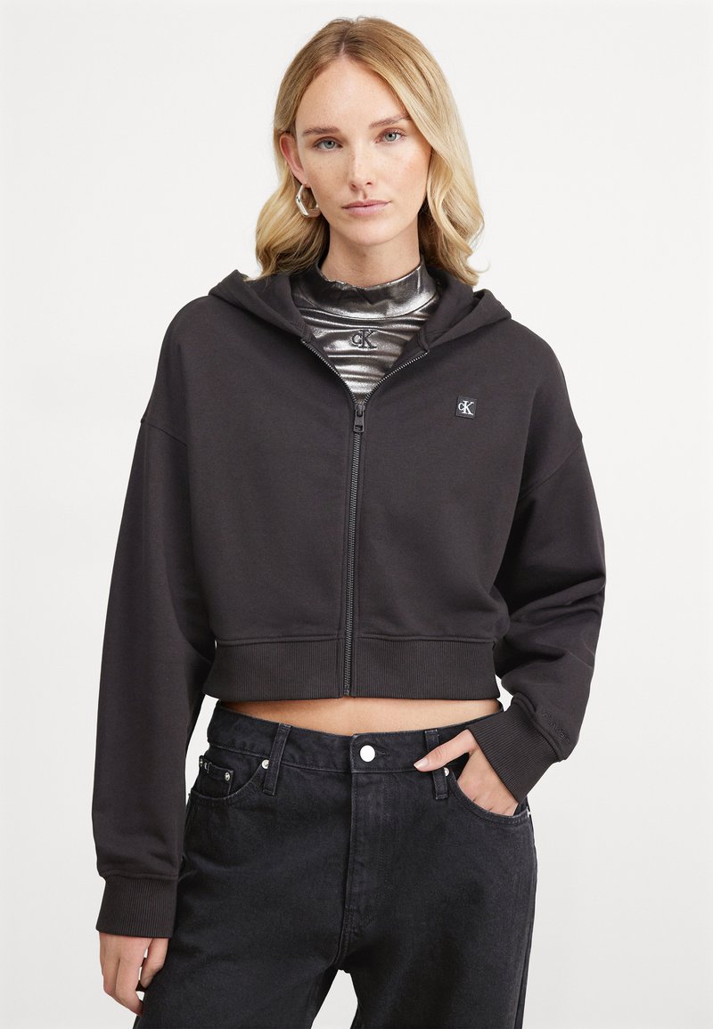 Hoodie noir cropped en tissu doux avec fermeture éclair et grande capuche. Présente un petit logo sur la poitrine et des poignets côtelés.