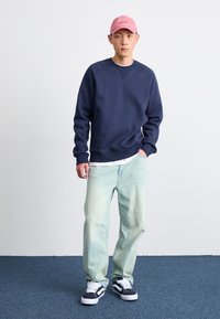 Marineblå sweatshirt, lyse denimbukser og sorte og hvide sneakers. Den lyserøde hat tilføjer et farvesplash. Enkel, afslappet pasform og casual stil.