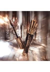 Schwarzer und goldener Mascara-Tube mit einem geometrischen, strahlenden Design. Begleitet von einem schwarzen Mascara-Bürstchen. Glänzende Oberfläche und deutlich beschriftet.