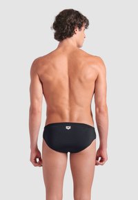 Junger Mann mit lockigem Haar, der schwarze Badebriefe trägt und mit dem Rücken zu einem schlichten grauen Hintergrund steht.
