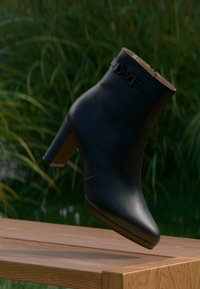 Bota de tornozelo em couro preto com bico alongado e salto bloco médio. Apresenta um detalhe decorativo em corrente e textura suave.