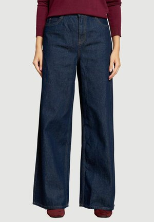 Szerokie, ciemnoniebieskie dżinsy z denimu noszone przez osobę ubraną w bordową bluzkę z długim rękawem i bordowe buty na jednolitym tle.