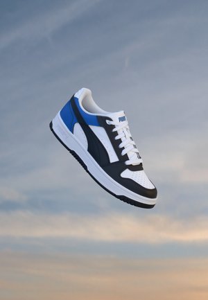 Basket Puma blanche, noire et bleue flottant devant un ciel doux et nuageux.