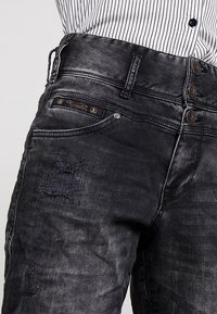 Svarta denimjeans med en sliten textur, femknapps gylf och en märkeslogotyp på midjan, med en smal passform.