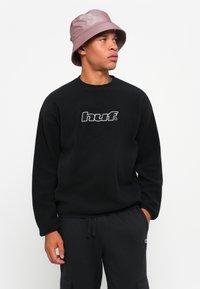 Sort fleece sweatshirt med "huf." logo, parret med sorte cargo-bukser. Modellen bærer en lys pink bøttehat. Simpelt, afslappet design.