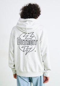Grå hettegenser med snøring som har en stor svart "Detroit" grafikk og lynbolt-design på baksiden. Laget av bomull.