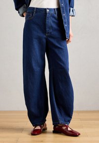 Μπλε τζιν παντελόνι φαρδιάς γραμμής με ψηλή μέση, διακοσμημένο με πορτοκαλί ραφές και μπροστινές τσέπες, συνδυασμένο με κόκκινα παπούτσια με λουστρίνι.