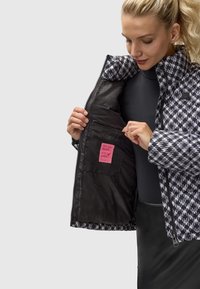 Padded jakna s crno-bijelim houndstooth uzorkom, zatvaranje na zip, i ružičasta oznaka unutarnje džepa. Glatka tekstura tkanine.