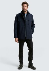 Tamsiai mėlynas vilnos mišinio švarkas tiesiu kirpimu, su dviem krūtinės kišenėmis, sagų užsegimu ir apykakle. Juodas tekstūruotas pamušalas.