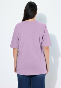 Übergrößen T-Shirt mit kurzen Ärmeln in lavendelfarbenem Baumwollstoff mit glatter Textur, Rundhalsausschnitt und lockerer Passform, kombiniert mit dunklen Jeans.