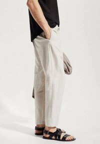 Pantalon en lin beige clair à coupe décontractée, associé à des sandales noires à lanières. La tenue comprend un tissu gris drapé sur un bras.