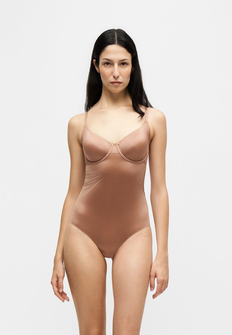 Femme aux longs cheveux foncés portant un body en satin marron clair ajusté avec de fines bretelles, debout devant un fond clair uni.
