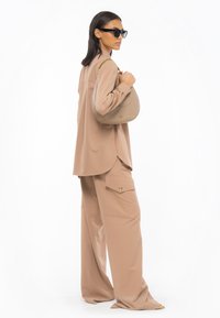 Blusa beige oversize con spacco laterale, abbinata a pantaloni larghi con tasche cargo. Gli accessori includono una borsa curva e occhiali da sole neri.