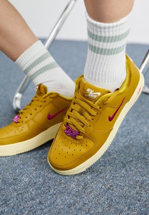 Żółte sneakersy Nike Air Force 1 z fioletowym znakiem Swoosh i ozdobami na sznurowadłach, noszone z białymi skarpetkami z zielonymi paskami, na niebieskim dywaniku.