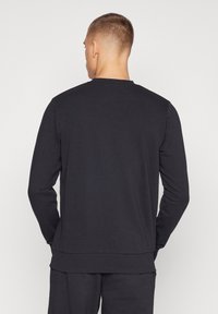 Homme aux cheveux courts, tourné de dos, portant un sweat-shirt noir uni à manches longues et un pantalon noir assorti.