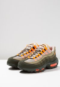 Pár sportovních tenisek v olivově zelené, béžové a oranžové barvě s síťovanými panely a viditelným vzduchovým odpružením na bílém povrchu.