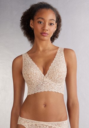 Béžový krajkový bralette s výstřihem do V, zvlněnými okraji a květinovými vzory. Měkká textilní struktura, širší ramínka a bezkosticový design pro pohodlí.
