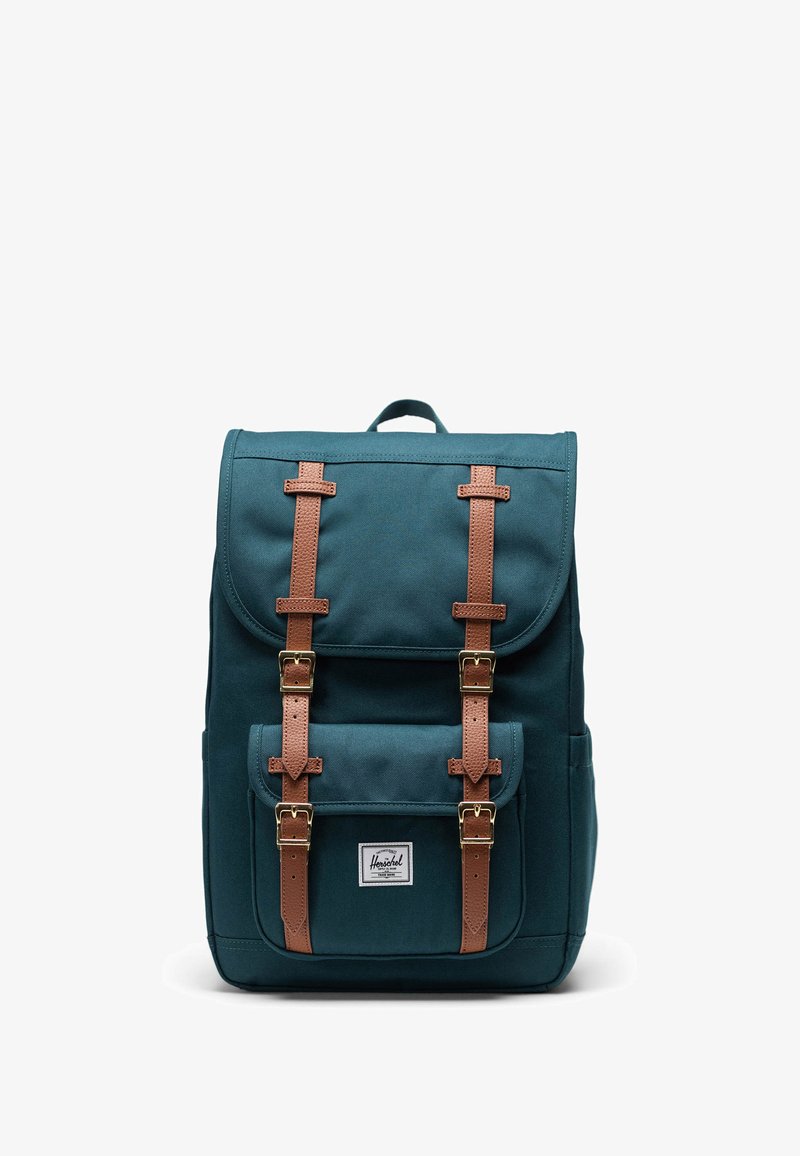 Turargrön canvasryggsäck med bruna läderremmar och detaljer, med en framficka och en logolapp på den nedre framsidan.