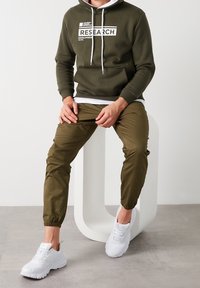 Sudadera verde oliva con texto blanco y cordones, combinada con pantalones jogger oliva y zapatillas blancas. Tejido suave, corte casual, diseño minimalista.