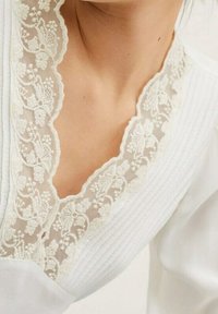 Hvid top med v-hals og intrikate blomsterer lace kanter. Stoffet er tekstureret med subtile lodrette linjer.